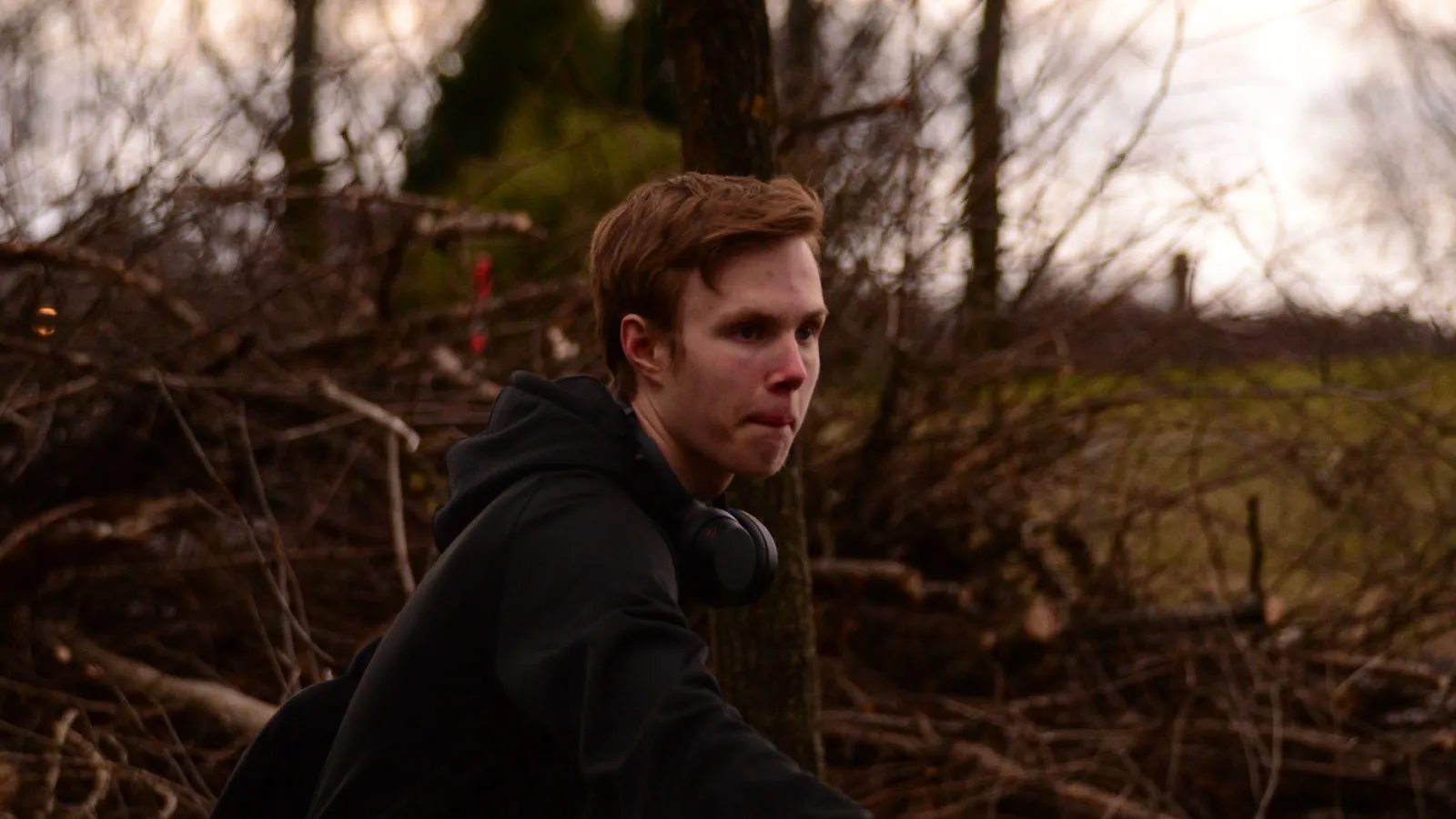 Een man in een donkere hoodie staat buiten tussen de bomen bij daglicht.