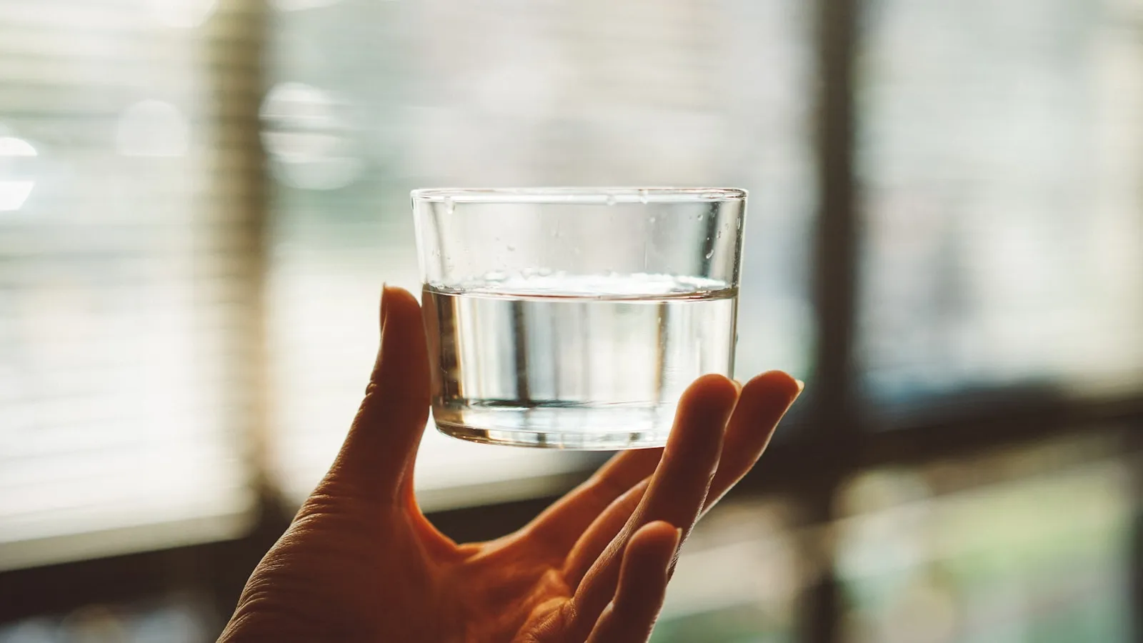 Hand houdt een helder glas half gevuld met water omhoog voor een raam met jaloezieen.