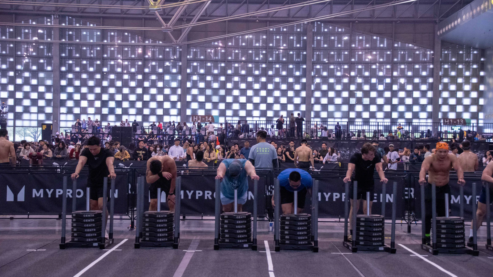 Deelnemers duwen de slee tijdens een Hyrox-wedstrijd in een volle evenementenhal.