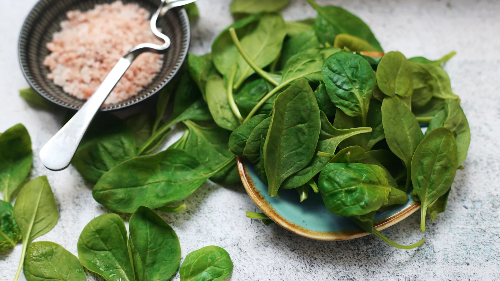 A bowl of fresh spinach next to a bowl of salt on a light table.