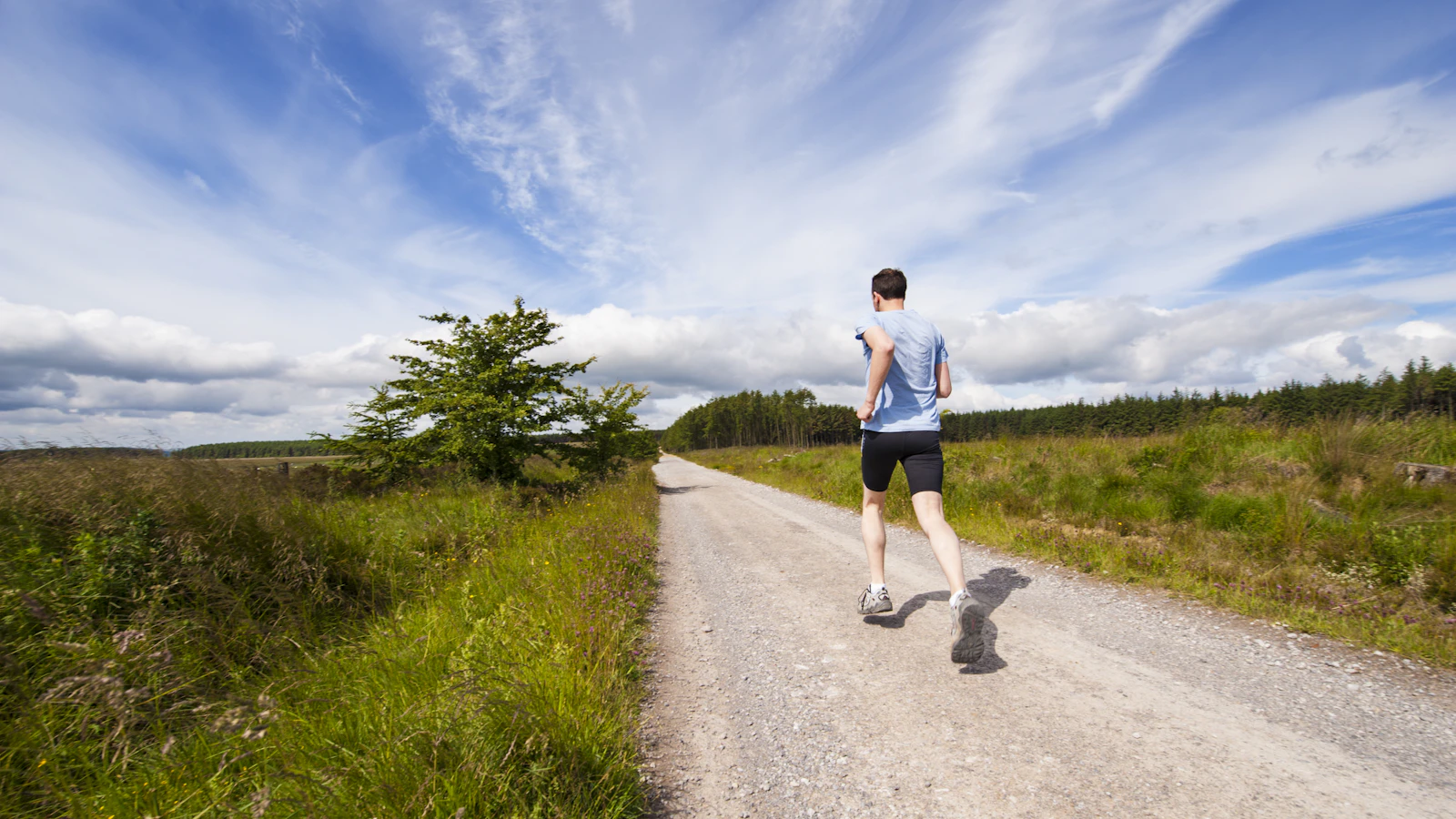 A man running on a gravel road between green fields under a blue sky.