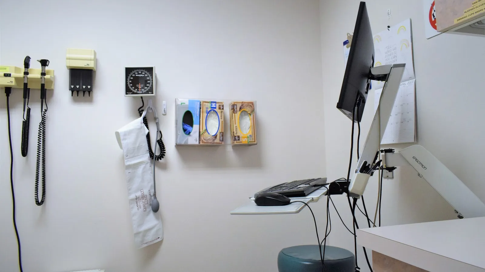 Lege onderzoekskamer met een bloeddrukmeter, otoscoop en handschoenendozen aan de muur.