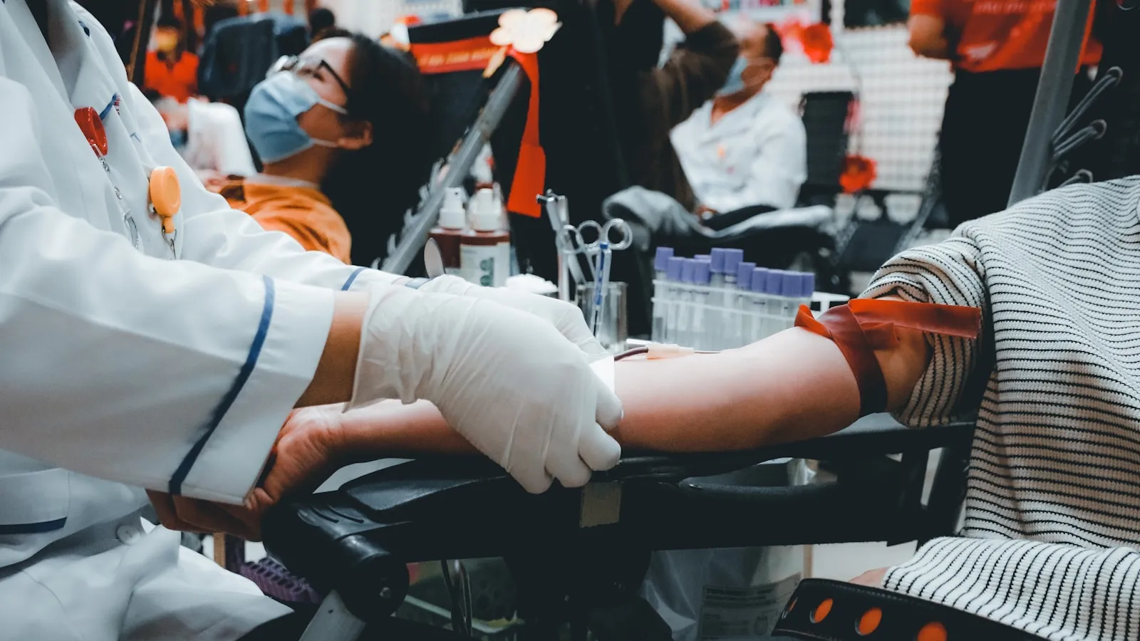 Zorgverlener met handschoenen neemt bloed af bij een arm met stuwband, met buisjes op de achtergrond.