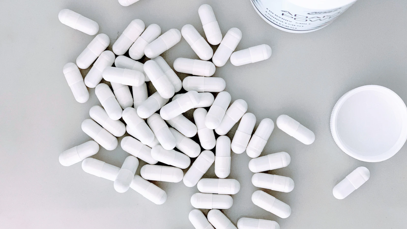 Witte capsules naast een geopend potje op een lichte ondergrond.