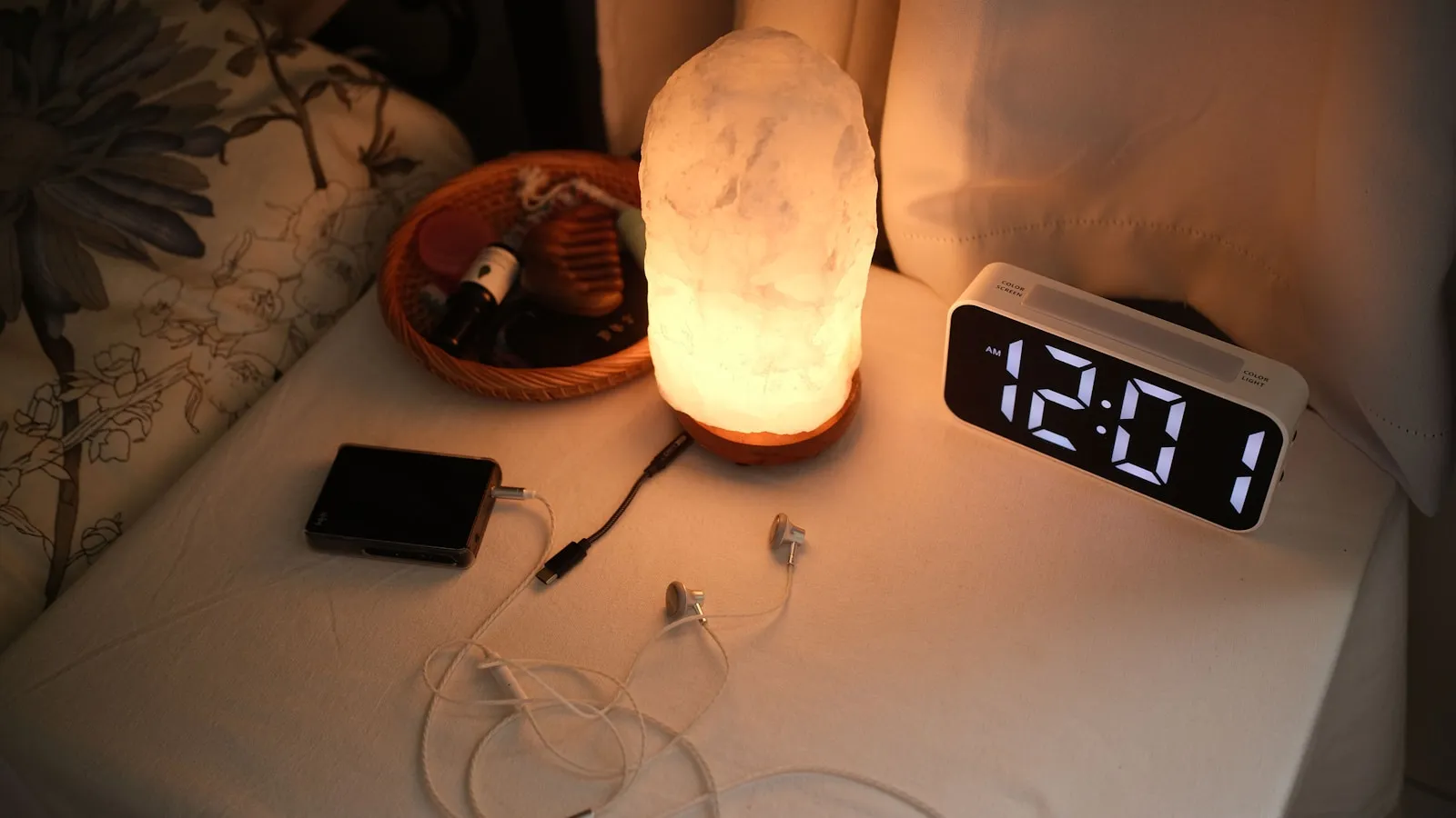 Een nachtkastje met een digitale wekker, een lampje en opladers naast het bed.