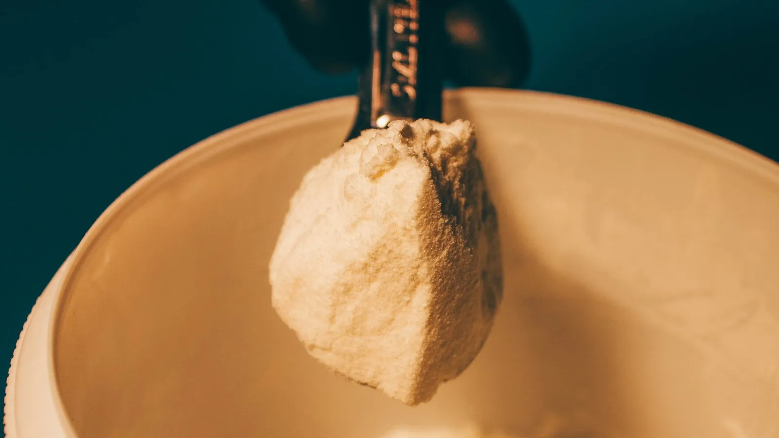 Een schepje wit supplementpoeder boven een pot, tegen een donkere achtergrond.
