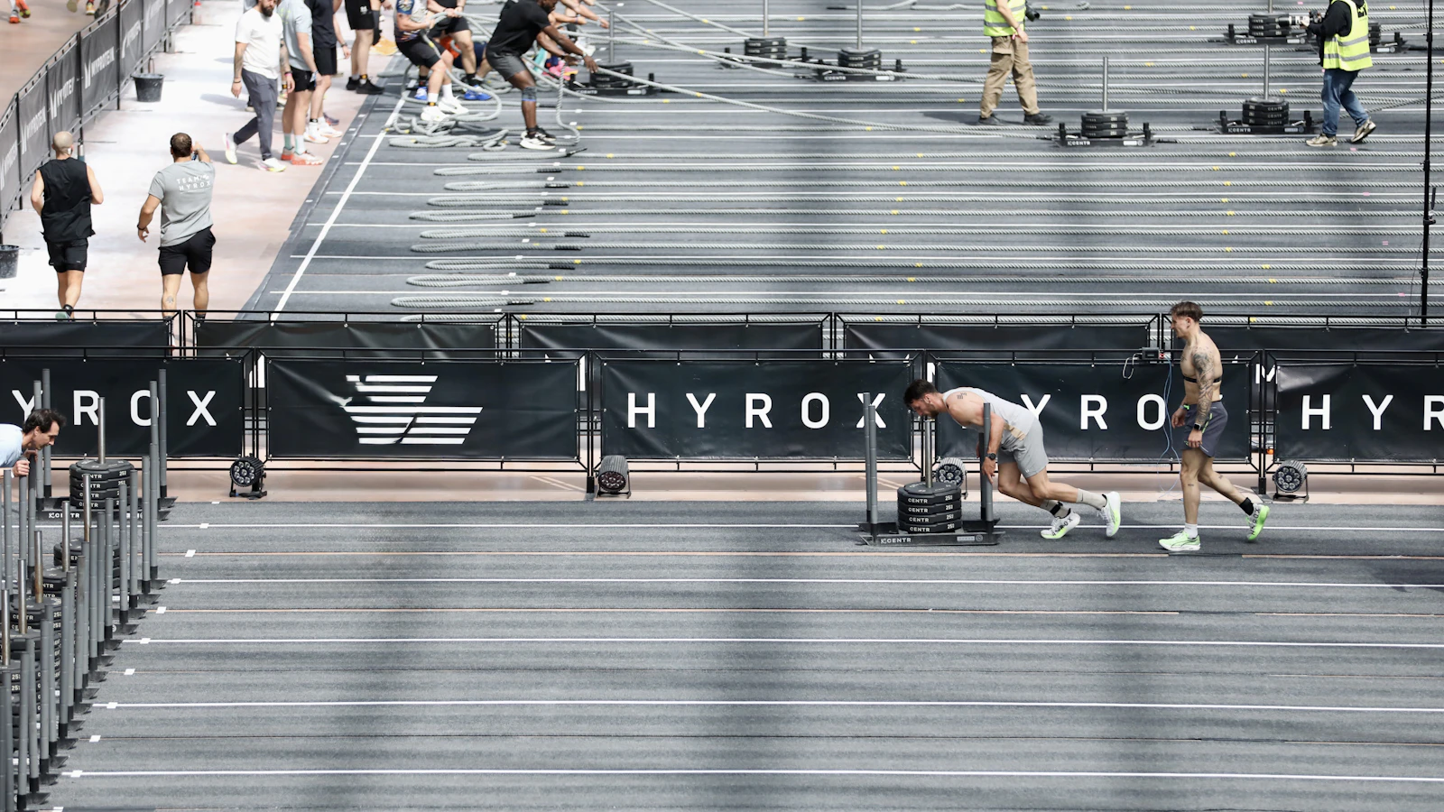 Overzicht van het Hyrox-parcours met stations en deelnemers in een evenementenhal.